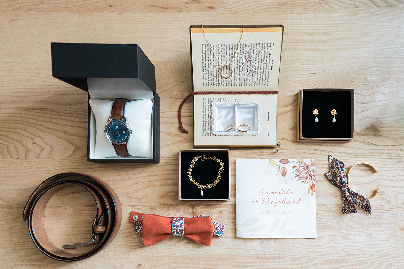 Ensemble d'accessoires mariages posés sur une table : faire-part, alliances, bijoux mariée, montre, ceinture et noeud papillon