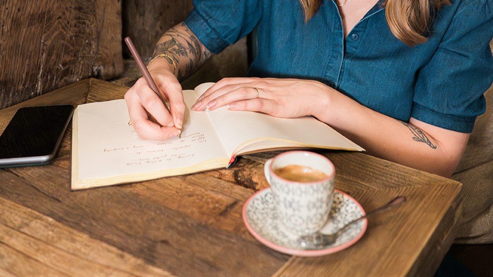 personne écrivant sur un carnet avec un café devant elle.